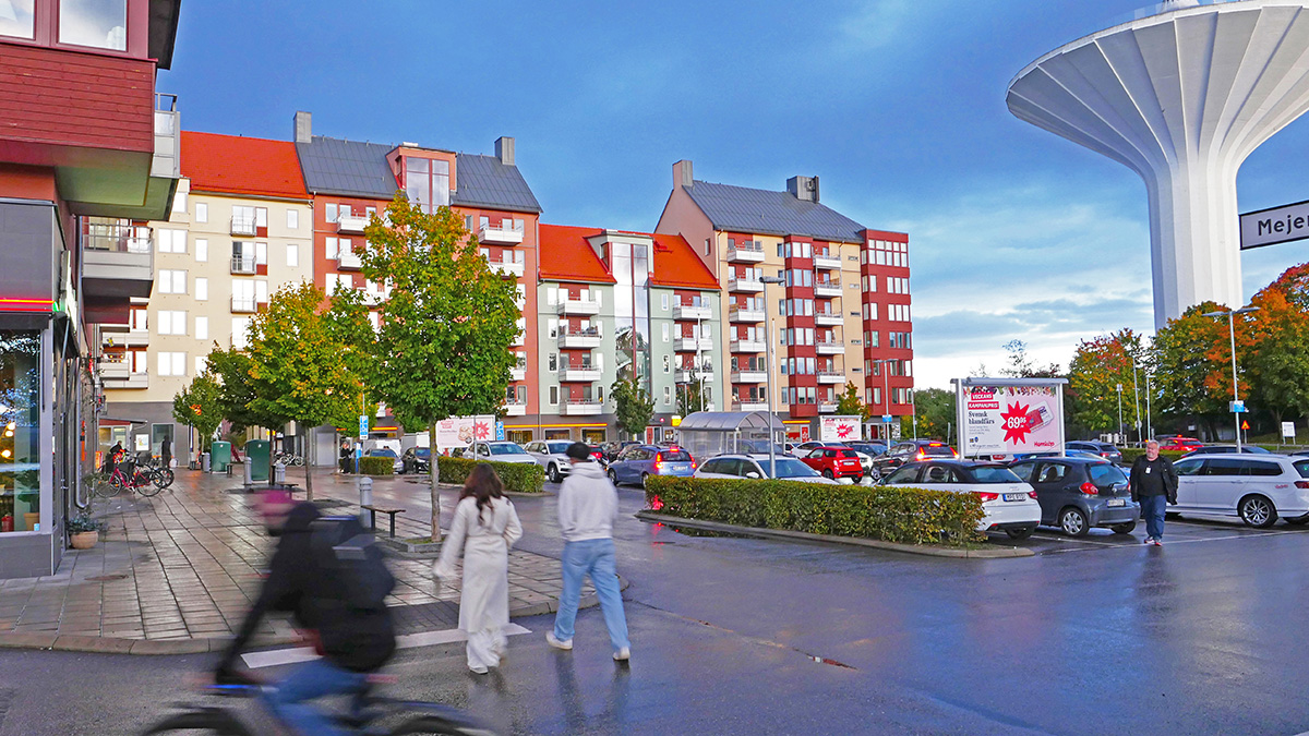 Stadsmiljö med flerbostadshus med röda tak, parkeringsplats med bilar och människor som går över gatan, samt ett stort vitt vattentorn i bakgrunden.