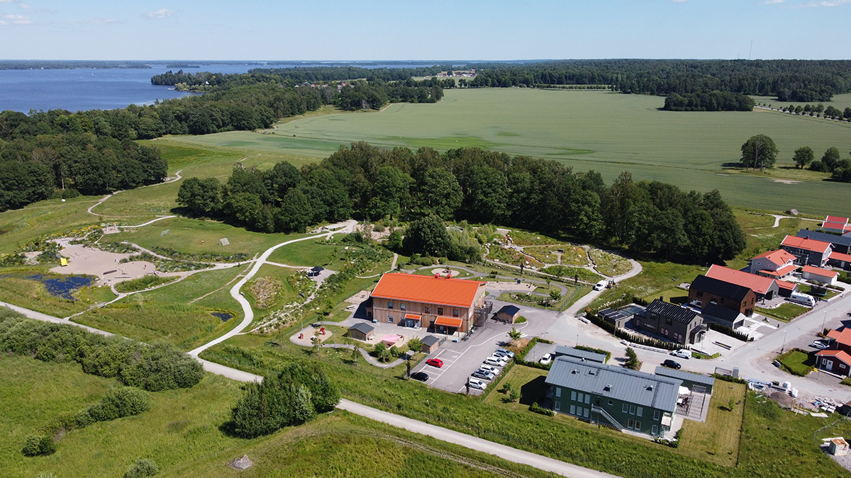 Flygbild över ett landskap med gröna fält, skog och en sjö i bakgrunden. I förgrunden syns en byggnad med rött tak, parkeringsplatser och gångvägar, omgiven av mindre hus och öppna grönområden.