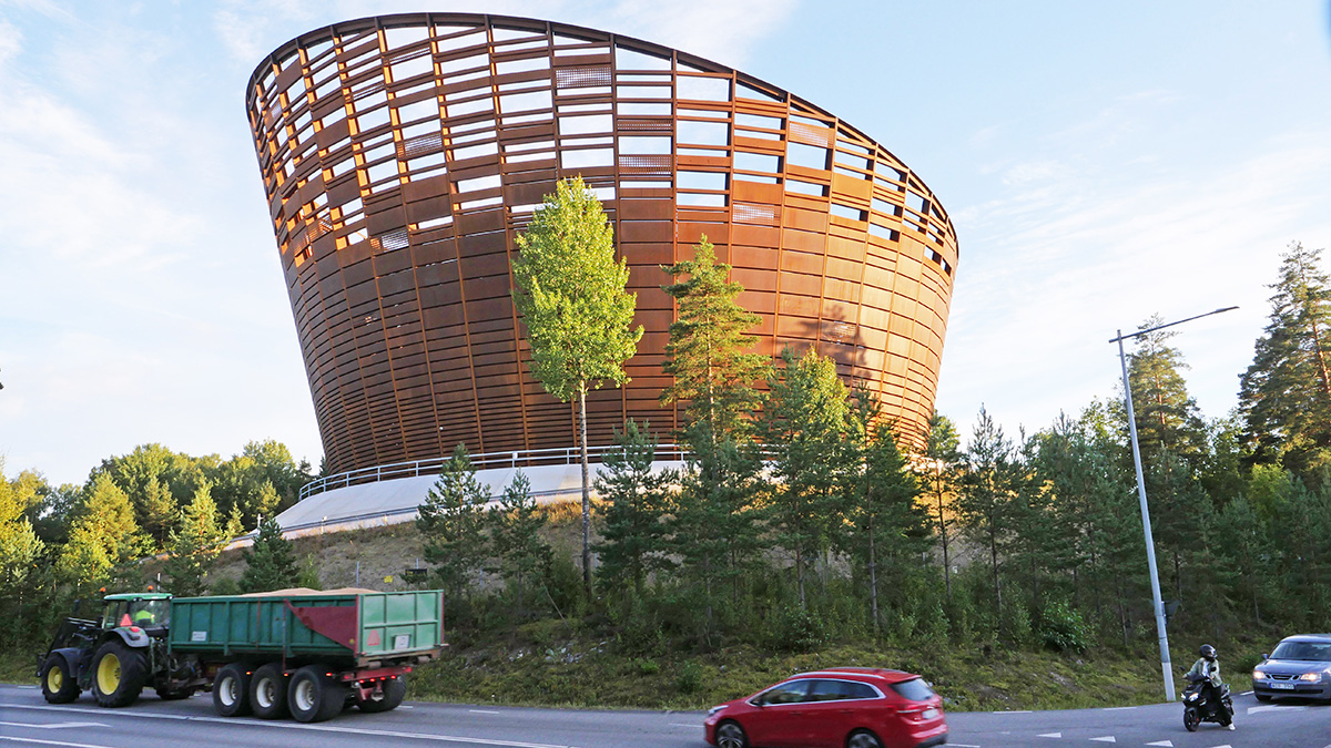 En stor, modern byggnad med böjd form och öppet rutmönster i rödbrunt trä, omgiven av höga tallar i en skogsmiljö under klar blå himmel.