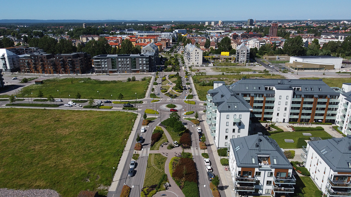Flygbild över ett modernt bostadsområde med flerfamiljshus, gröna ytor och en bred gata med rondeller och planteringar. I bakgrunden syns stadens centrum med fler byggnader.