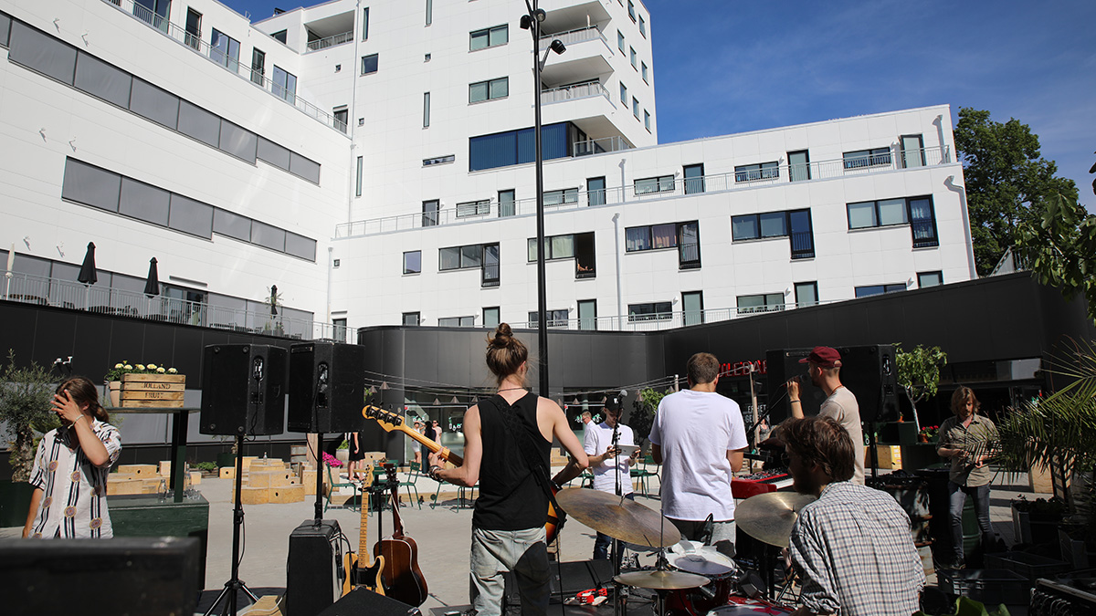 Utomhusscen med ett band som spelar musik framför en modern vit byggnad med balkonger och stora fönster. Publik och musikinstrument syns i förgrunden.