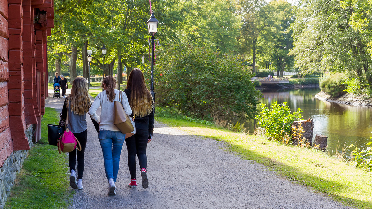 Tre personer promenerar längs en grusgång vid ett vattendrag, omgiven av grönska och träd, med rödmålade byggnader på ena sidan.