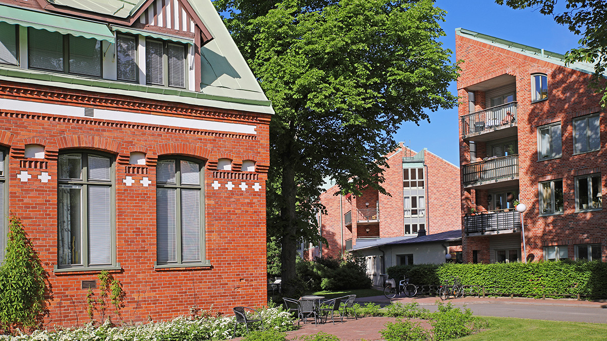 Bostadsområde med tegelhus och moderna flerfamiljshus, omgiven av träd och grönska under en klar blå himmel.