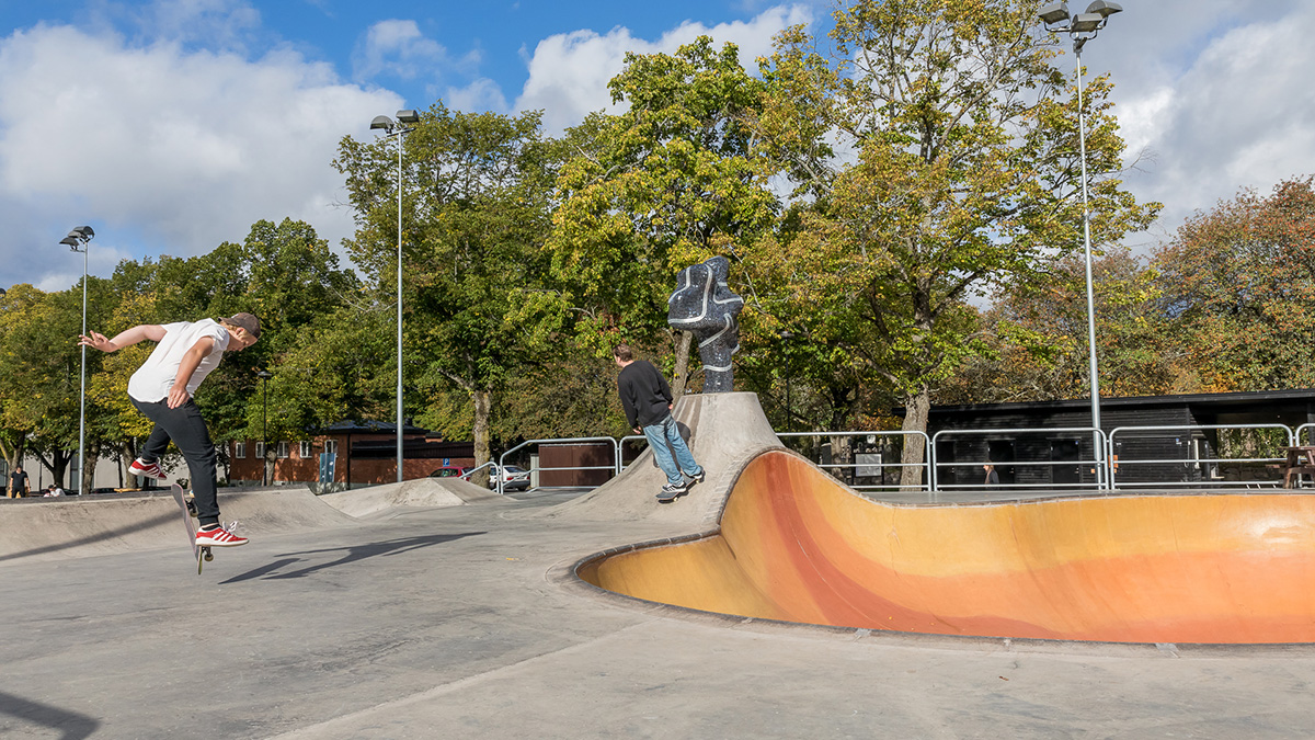 Skatepark med två personer som åker skateboard vid en färgad betongramp, omgiven av träd och en staty i mitten.