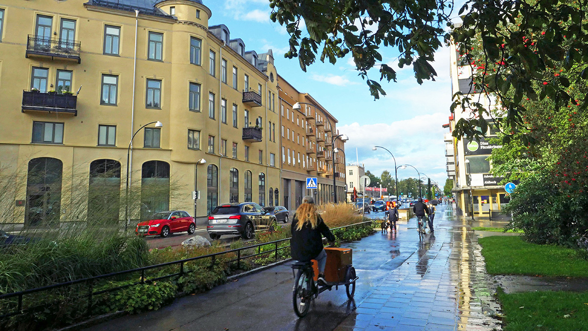 Stadsgata med cyklister och fotgängare längs en trottoar efter regn, med äldre byggnader och parkerade bilar på ena sidan och grönska på den andra.