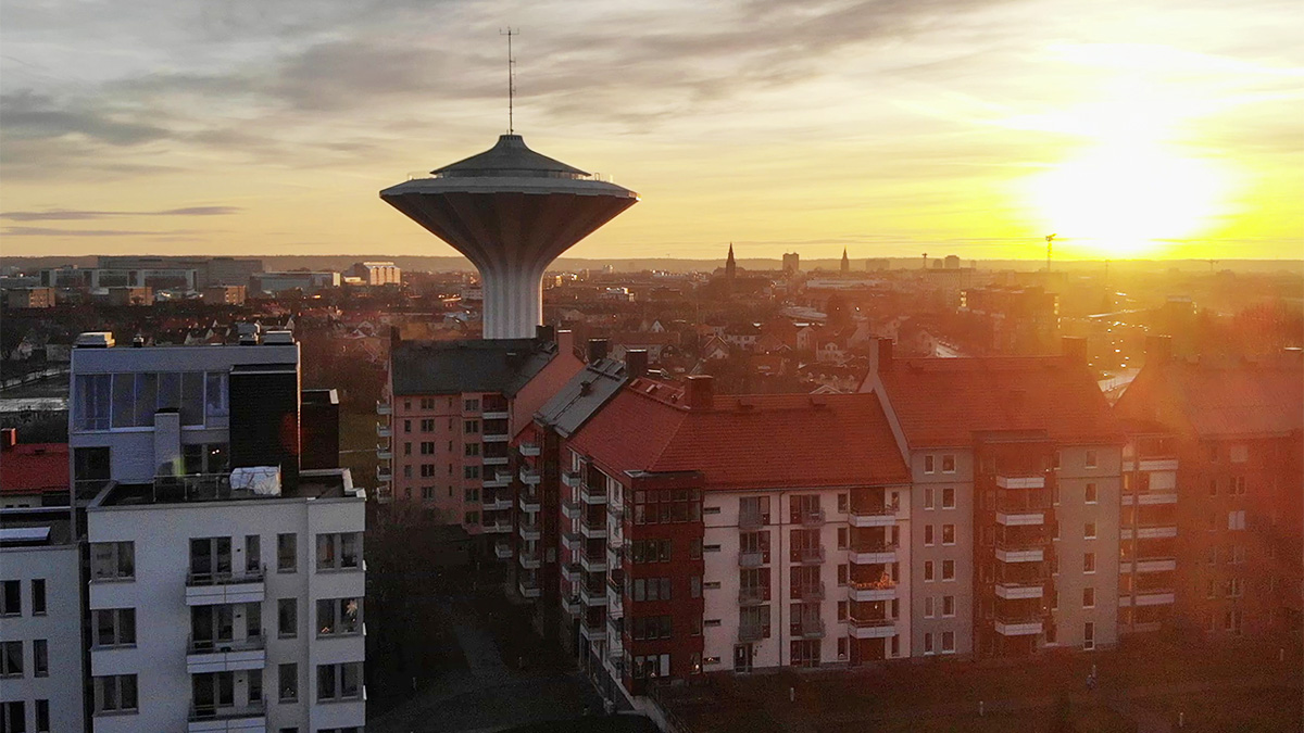 Solnedgång över Örebro med vattentornet Svampen i centrum, omgiven av flerbostadshus med röda tak och stadens silhuett i bakgrunden.