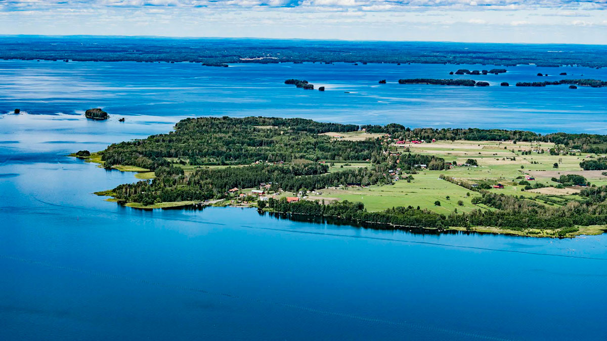 En flygbild av Vinön, en stor ö omgiven av sjön Hjälmaren. Ön är täckt med grönskande skog och öppna fält, med några spridda byggnader och vägar synliga. I bakgrunden syns fler mindre öar och en vidsträckt horisont under en himmel med lätta moln.