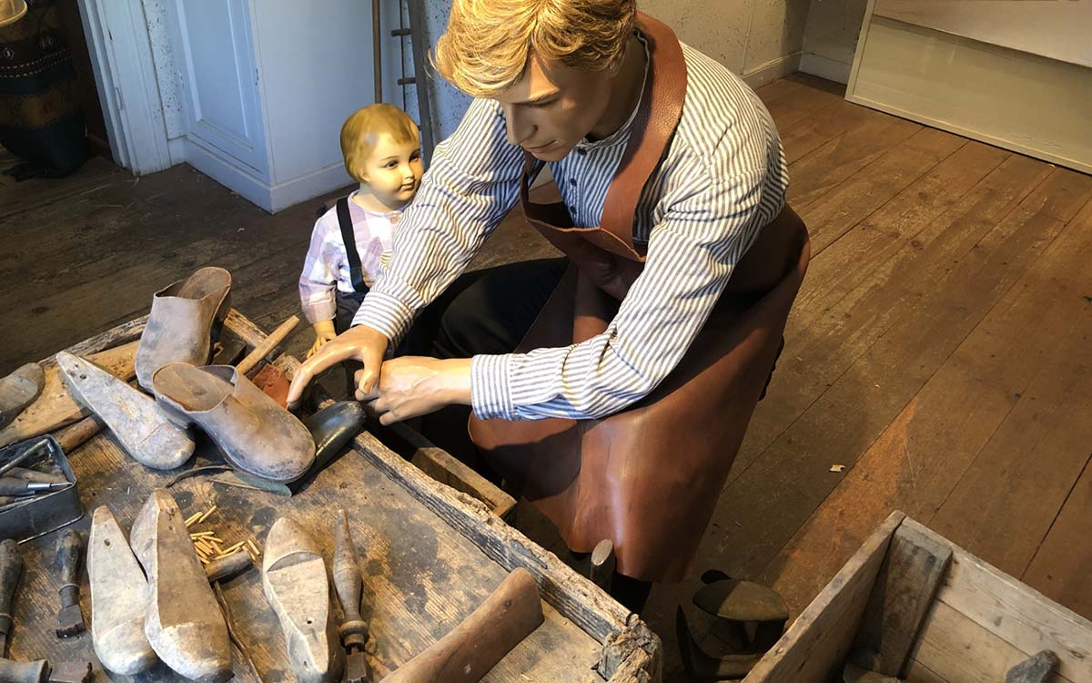 Bild på interiör från skomakerimuseet och skomakare samt ett barn som står och tittar på när skomakaren lagar ett par träskor