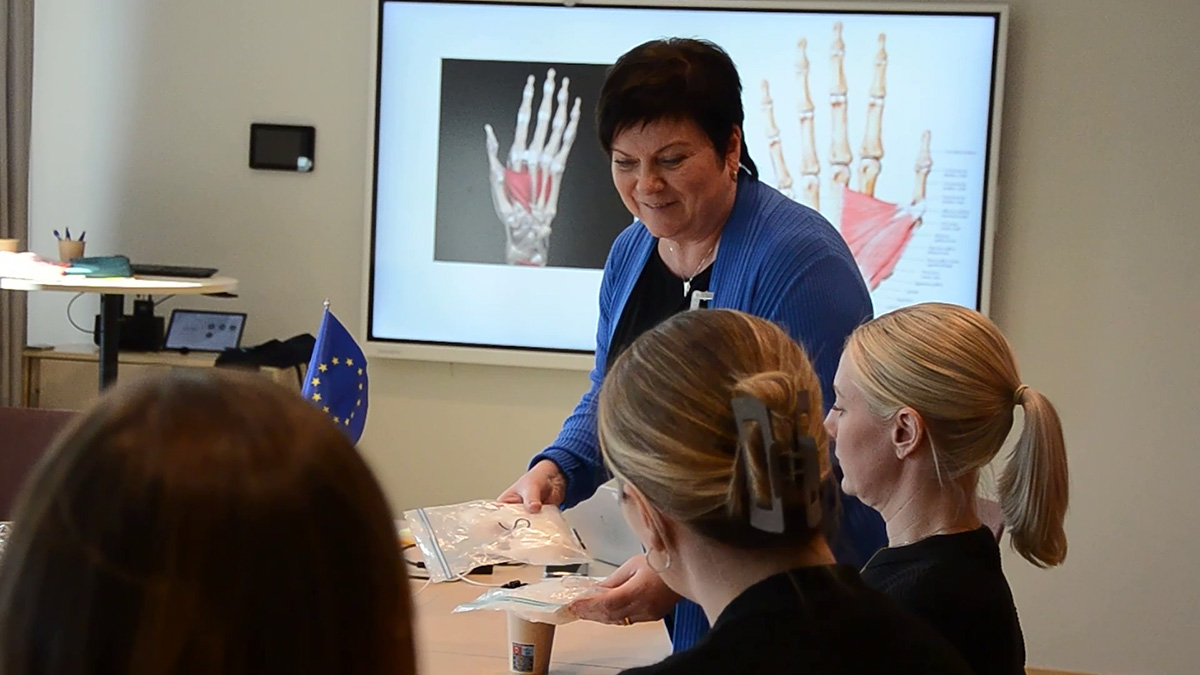 En grupp människor befinner sig i ett mötesrum. En person står upp och håller i material, medan tre andra sitter och lyssnar. I bakgrunden visas en anatomisk bild av en hand med muskler och ben på en stor skärm. På bordet ligger en EU-flagga.
