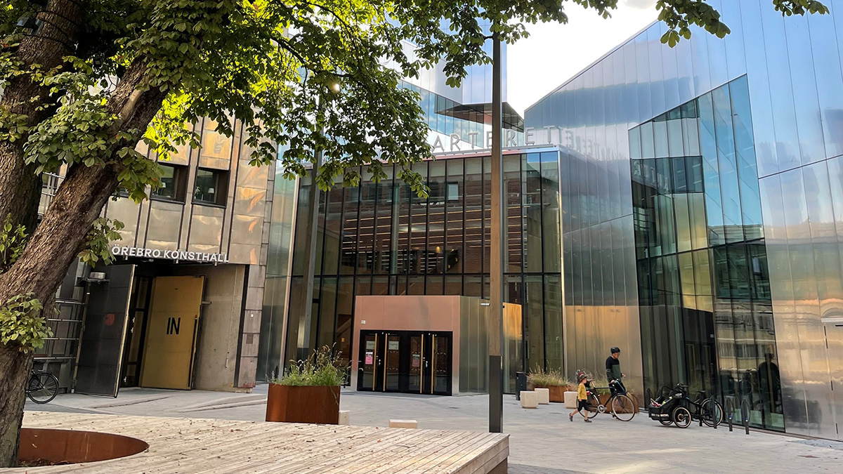 Modern byggnad med glasfasader och metallpaneler i ett öppet torgområde. Till vänster syns en ingång med texten 'Örebro Konsthall', och i mitten en större entré med glasdörrar. Framför byggnaden står en cykel och en person med barn på cykel passerar. Ett stort träd med gröna blad skymtar i förgrunden.