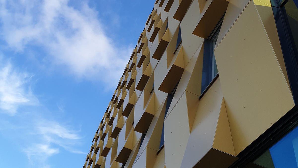 Fasad på en modern byggnad med guldfärgade, geometriska paneler som skapar ett mönster av utskjutande former. Några fönster är synliga mellan panelerna, och himlen i bakgrunden är blå med tunna vita moln.
