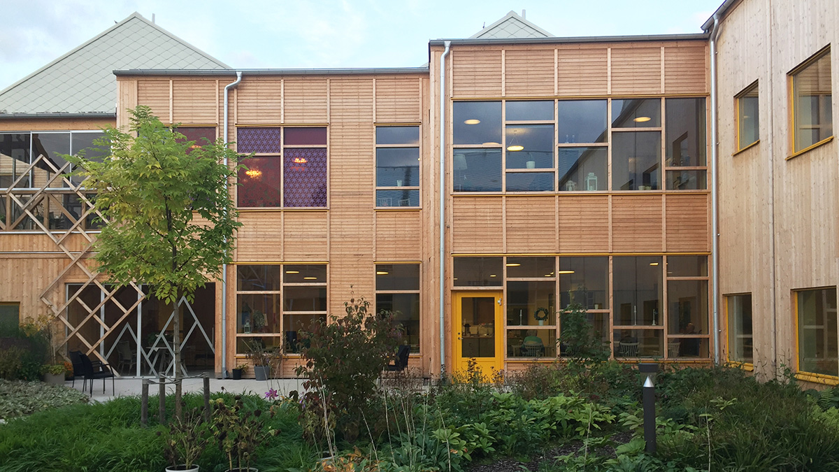 Fasad på en modern byggnad i trä med stora fönster och guldfärgade detaljer runt karmarna. Byggnaden har två våningar och omges av en grön trädgård med buskar och ett mindre träd i förgrunden. Ljuset är mjukt och himlen är delvis molnig.
