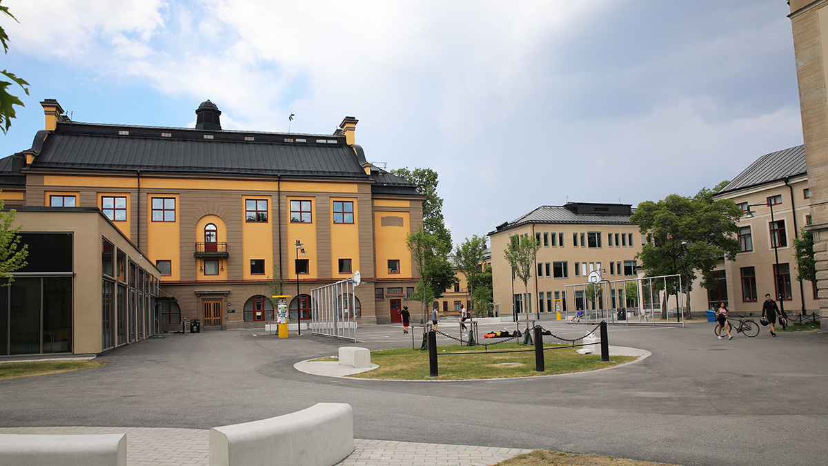 Öppen skolgård med moderna och äldre byggnader i ljusa färger. Den största byggnaden har gul fasad med svarta takdetaljer och flera fönster. I mitten finns en rund yta med bänkar och stolpar, och några personer syns gående eller cyklande. Himlen är delvis molnig.