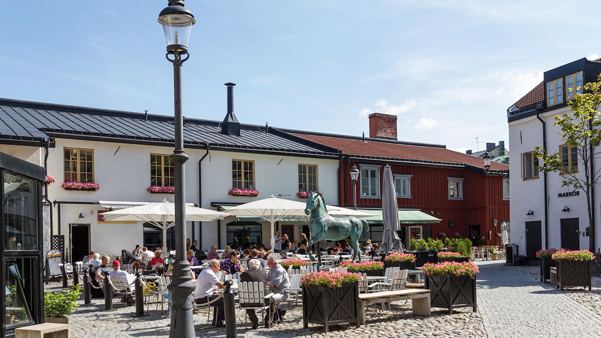 Solig torgmiljö med uteserveringar och människor som sitter vid bord under parasoller. Torget är omgivet av äldre byggnader med vita och röda fasader, svarta tak och blomlådor under fönstren. I mitten står en grön hästskulptur och flera blomkrukor med rosa blommor. En gatlykta syns i förgrunden.