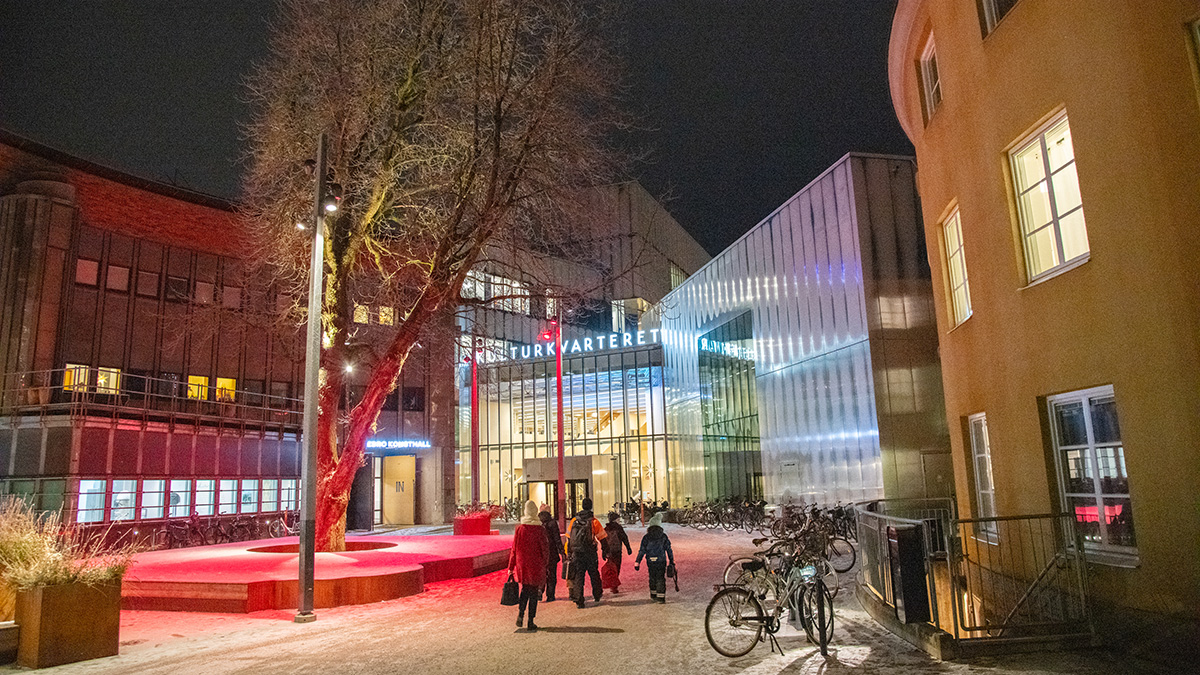 Kvällsbild av Kulturkvarteret med upplysta fasader och glasentré. Ett träd med röd belysning står i mitten av torget, och flera personer promenerar mot ingången. Cyklar är parkerade längs byggnaden.