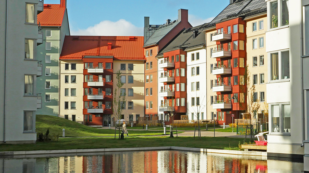 Moderna flerfamiljshus med ljusa fasader och röda tak vid en spegelblank vattenyta. Husen har balkonger och stora fönster, och i bakgrunden syns en grön gräsyta med gångvägar och några människor som promenerar.