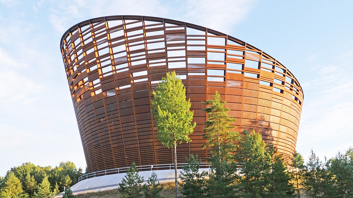 Stort, konformat byggnadsverk med rostfärgad metallstruktur och öppna paneler, omgivet av gröna träd och blå himmel i bakgrunden.
