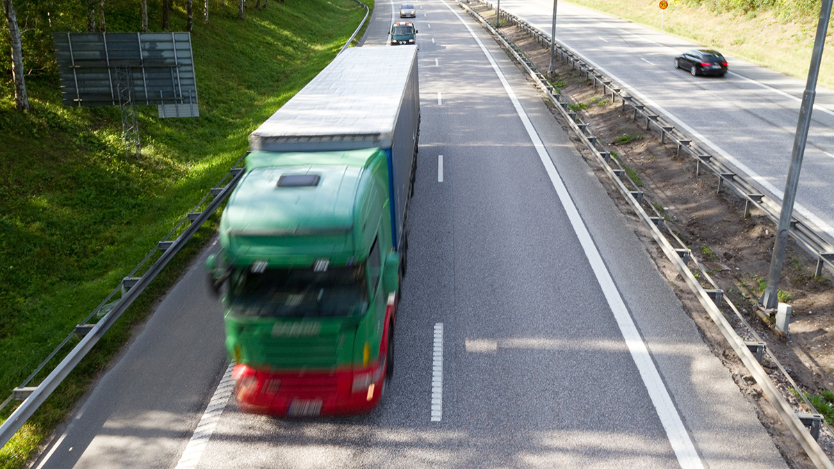 En grön lastbil med röd front kör på en motorväg i dagsljus, fotograferad uppifrån. På höger sida syns en parallell väg med en svart bil och flera skyddsräcken. Bakgrunden består av grön grässluttning och några träd.