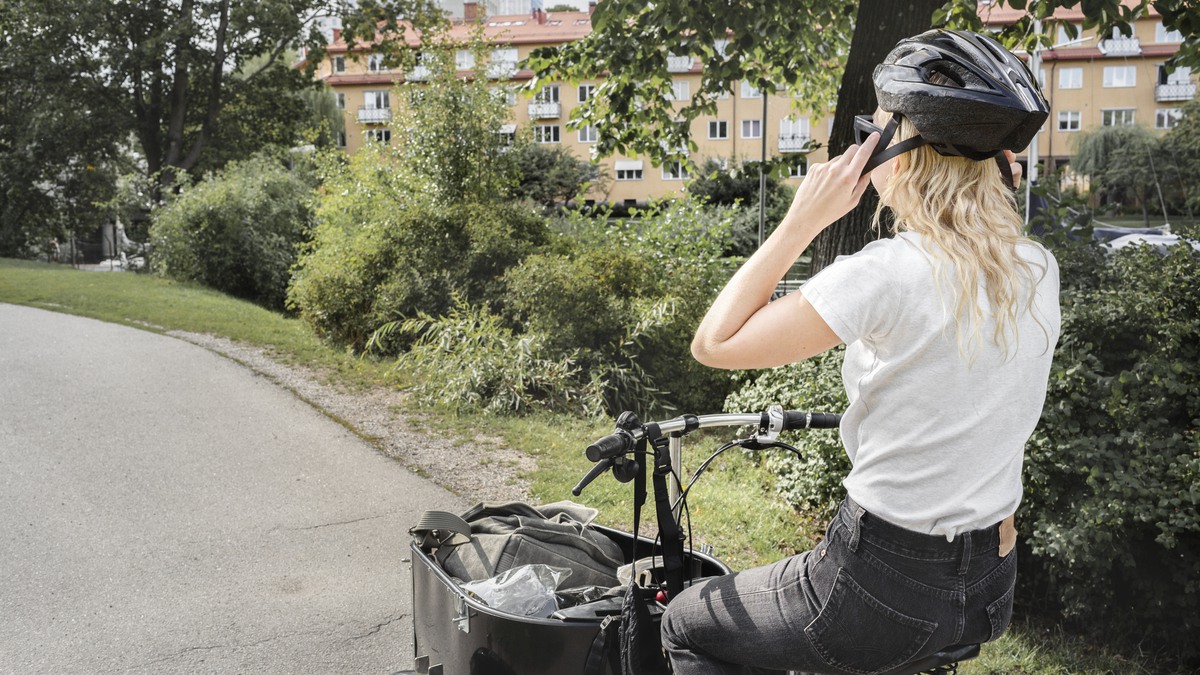En person med långt, blont hår sitter på en trehjulig cykel och justerar sin svarta hjälm. Personen bär en vit t-shirt och mörka byxor. I bakgrunden syns gröna buskar, träd och ett gult flerfamiljshus med röda tak. Cykeln har en stor lastkorg framtill. Stigen personen cyklar på är asfalterad och slingrar sig genom ett grönområde.