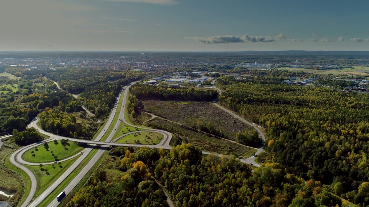 Flygfoto av ett landskap med en motorvägskorsning omgiven av frodiga gröna skogar och fält. Motorvägen böjer sig och korsar en annan väg, vilket skapar ett klöverbladsmönster. I bakgrunden syns en stad med många byggnader och strukturer som sträcker sig mot horisonten under en klarblå himmel med spridda moln.