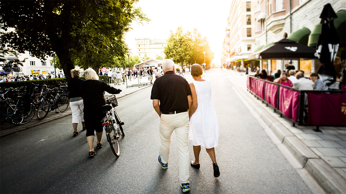 Ett par går hand i hand längs en solbelyst gata kantad av träd och byggnader. Mannen bär en svart skjorta och beige byxor, medan kvinnan är klädd i en vit klänning. Till vänster om dem skjuter en annan kvinna en cykel. På höger sida av gatan sitter människor på ett utomhuscafé med rosa avspärrningar. I bakgrunden syns fler fotgängare och cyklister som njuter av den soliga dagen.