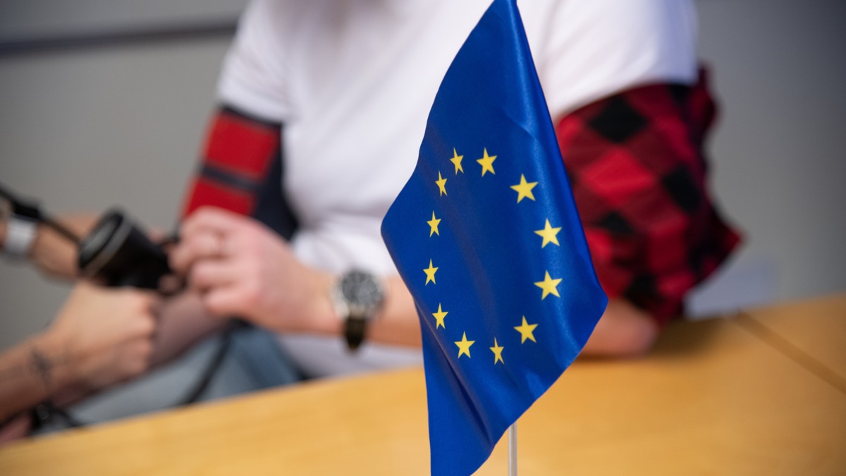 Ett litet bord placerat framför en suddig bakgrund där en person får sitt blodtryck mätt. I förgrunden står en liten EU-flagga som är i tydlig fokus.