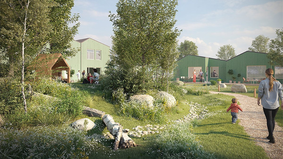 Lummig utomhusmiljö med grusgångar, stenar och grönska framför flera låga, gröna byggnader. Flera personer syns utspridda i området, bland annat en vuxen som går på en stig tillsammans med ett barn. Några personer sitter eller står nära byggnaderna och ett öppet träskärmtak. Soligt väder och mycket vegetation omger platsen.