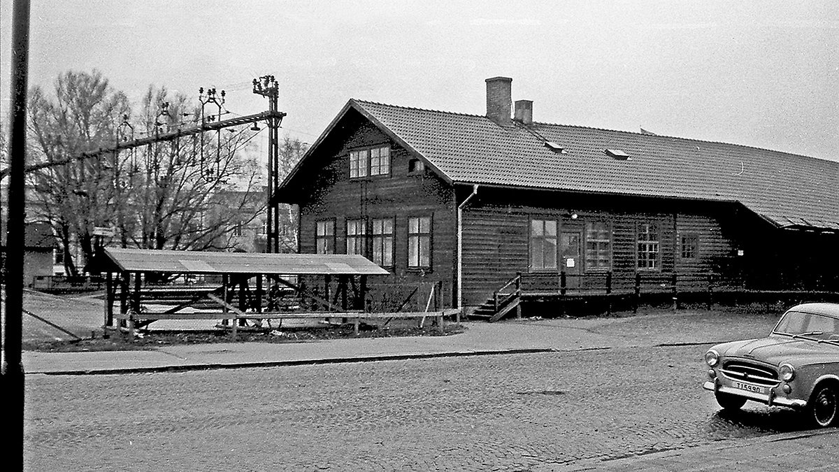 Historisk svartvit fotografi av en låg träbyggnad vid ett järnvägsområde, med sadeltak, skorsten och flera fönster. Intill byggnaden finns ett enkelt perrongtak och stolpar för kontaktledning. I förgrunden syns en kullerstensgata där en äldre personbil står parkerad. Träd och ytterligare byggnader finns i bakgrunden.